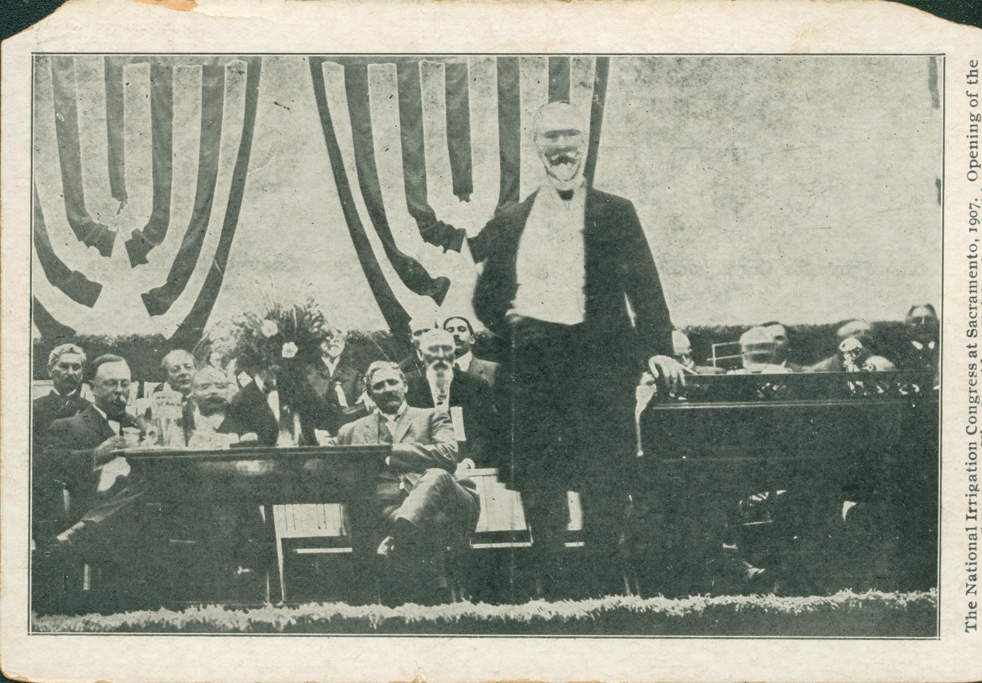 This postcard shows a Mr. Fairbanks giving a speech with his back to several men seated at desks.
